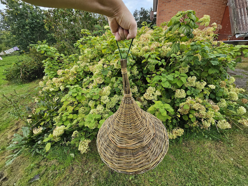 Handwoven willow bird feeder held up by its hanging cord in front of a hydrangea bush, showing how the cone-shaped roof hangs