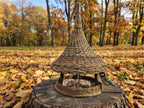 Tall handwoven willow bird feeder with a bell-shaped roof and spire top on a tree stump among autumn leaves, a great tit at the seed tray