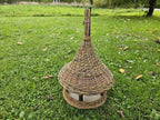 Tall handwoven bird feeder with a spire top standing on the grass in an orchard, seen from a low angle