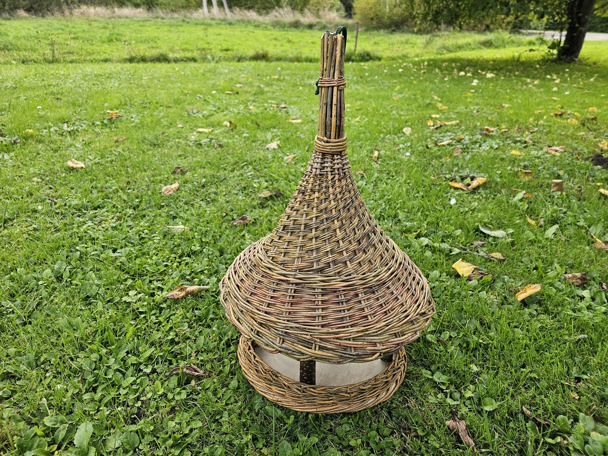Tall handwoven bird feeder with a spire top standing on the grass in an orchard, seen from a low angle