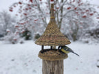 Tall willow bird feeder with bell roof set on a wooden post in a snowy garden, a great tit eating from the platform