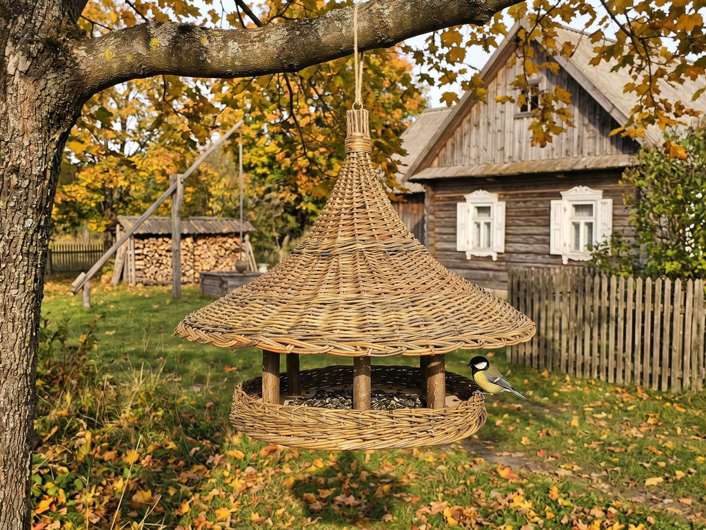 Large wicker bird feeder with a wide woven roof hanging from an apple tree in an autumn garden, with a great tit on the seed platform