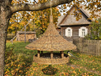 Large wicker bird feeder with a wide woven roof hanging from an apple tree in an autumn garden, with a great tit on the seed platform