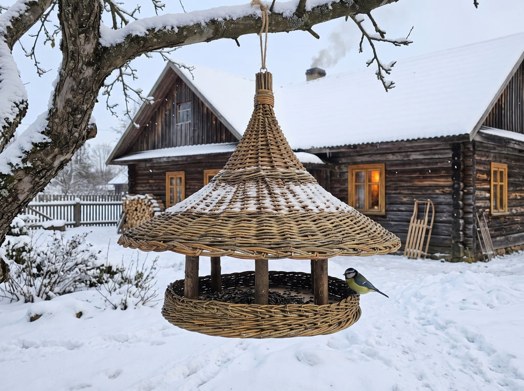 Handwoven willow bird feeder hanging from a snowy branch in front of a wooden farmhouse in winter, with a blue tit at the platform