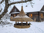 Handwoven willow bird feeder hanging from a snowy branch in front of a wooden farmhouse in winter, with a blue tit at the platform