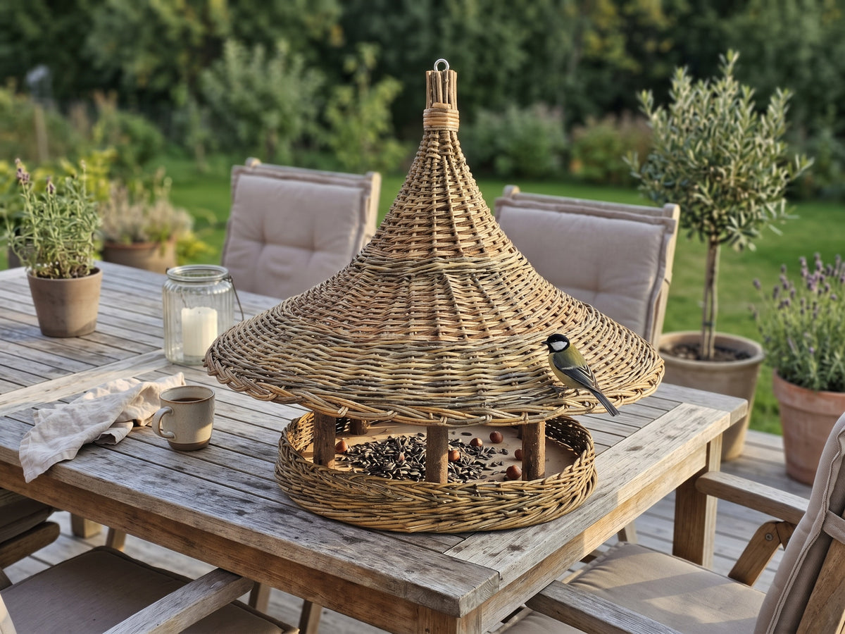 Willow bird feeder with a wide woven brim on a wooden patio dining table, with a great tit perched on the roof edge