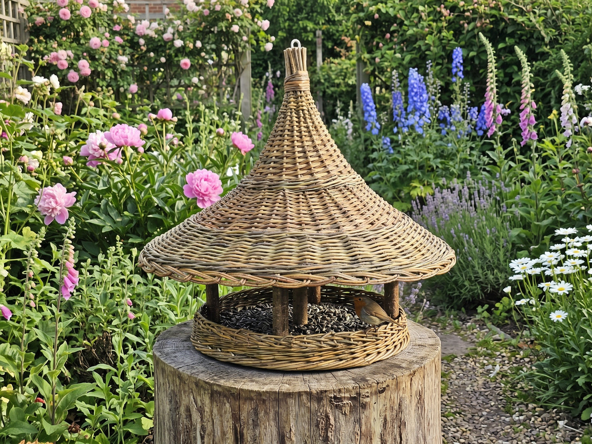 Extra large wicker pagoda feeder filled with seed on a tree stump in a flower garden with peonies, delphiniums and foxgloves