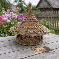 Extra large handwoven willow pagoda bird feeder with a tall pointed roof on a rustic garden table, a sparrow pecking seed beside it
