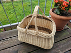 Handwoven willow garden basket with two crossed handles and braided edge on a wooden balcony table beside a pot of flowers.