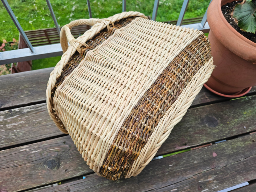 Handwoven two-handled willow basket tipped on its side on a balcony table, showing the shape of the body and braided edge.