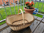 Large oval willow shopping basket with braided rim and tall handle on a balcony table beside a pot of flowers