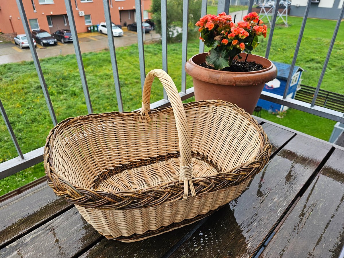 Large oval willow shopping basket with braided rim and tall handle on a balcony table beside a pot of flowers