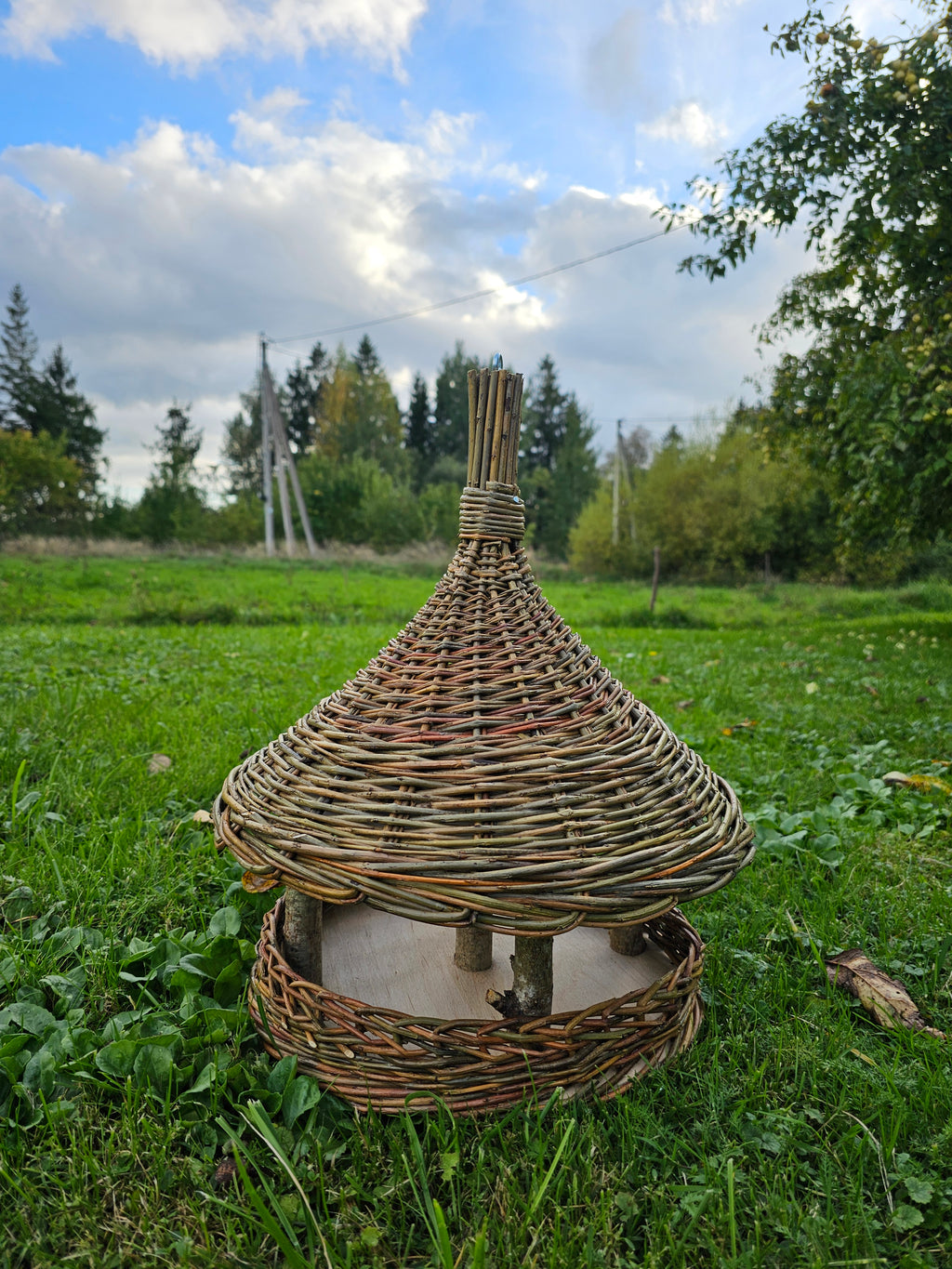 Wicker bird feeder with a tall woven spire standing on the grass in an open meadow, showing the cone roof, wooden posts and round seed tray.