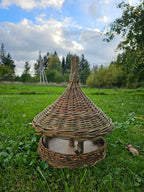 Wicker bird feeder with a tall woven spire standing on the grass in an open meadow, showing the cone roof, wooden posts and round seed tray.