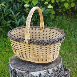 Ovaler Sammelkorb aus Weide mit Zopfrand und kräftigem Bügelhenkel auf einem Birkenstumpf im grünen Garten