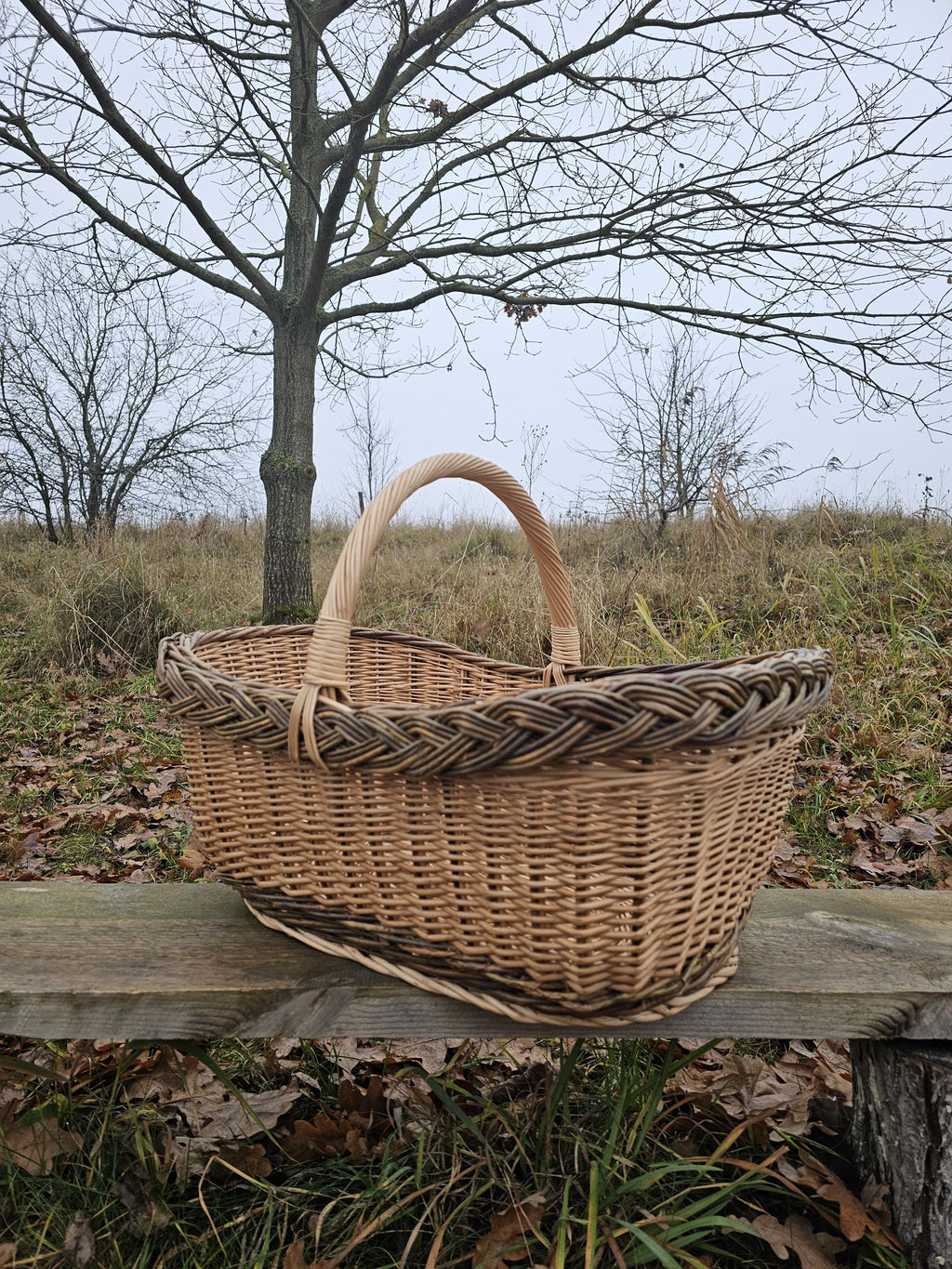 Side view of the large oval wicker market basket with a tall handle and braided rim on a wooden bench in a meadow