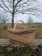 Side view of the large oval wicker market basket with a tall handle and braided rim on a wooden bench in a meadow