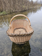 Oval willow harvest basket standing in clear lake shallows, reeds behind it and the woven base visible through the water.