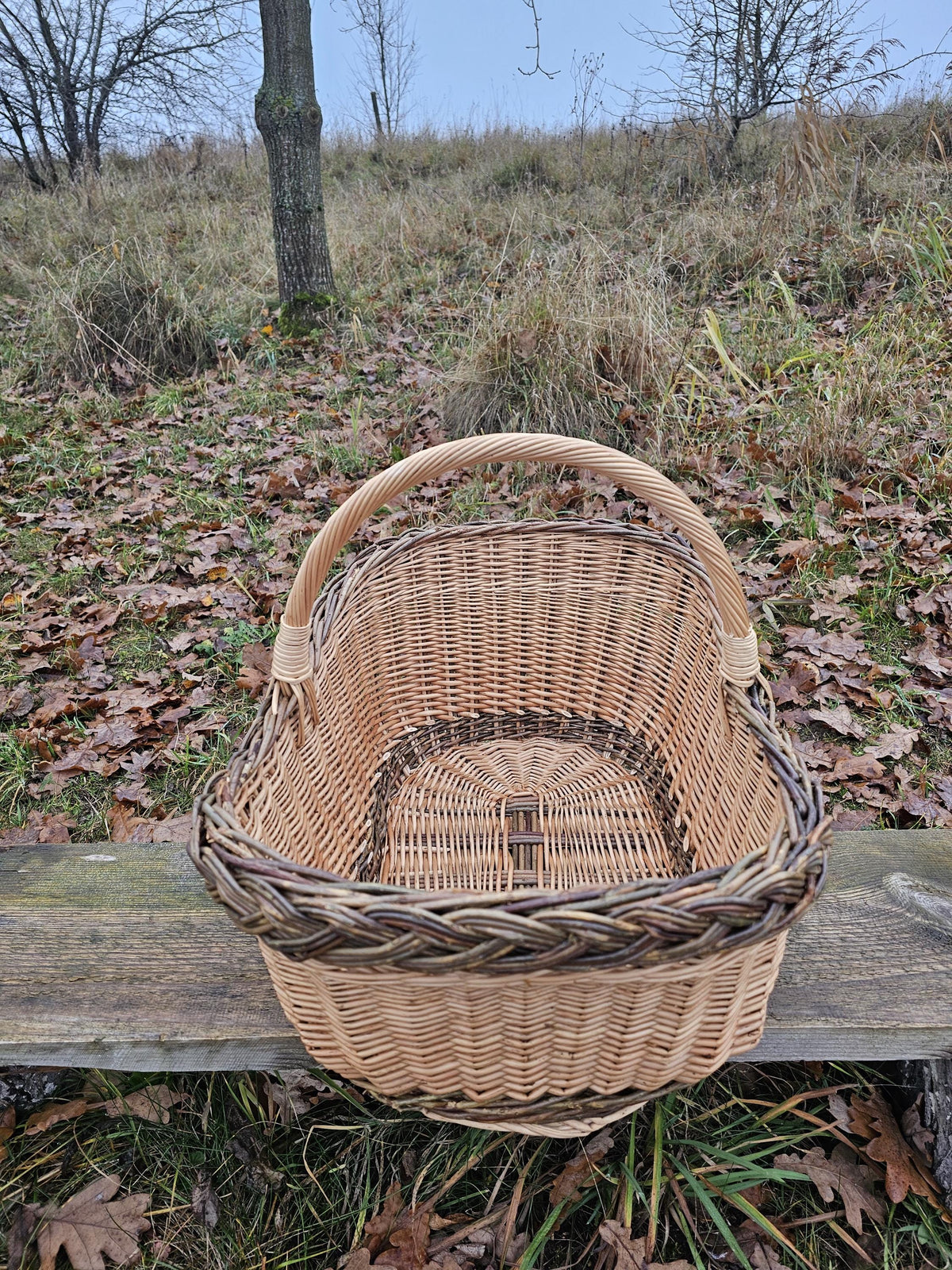 Top view of the large oval willow basket with handle on a wooden bench, showing the woven base and braided rim