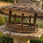 Close-up under the roof of the willow bird feeder: bark-on branch posts, wooden roof lining and deep woven rim, on a stone garden pedestal
