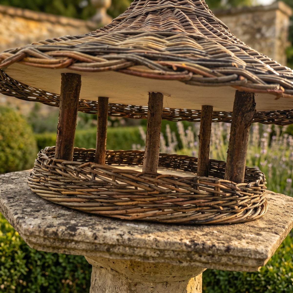 Close-up under the roof of the willow bird feeder: bark-on branch posts, wooden roof lining and deep woven rim, on a stone garden pedestal