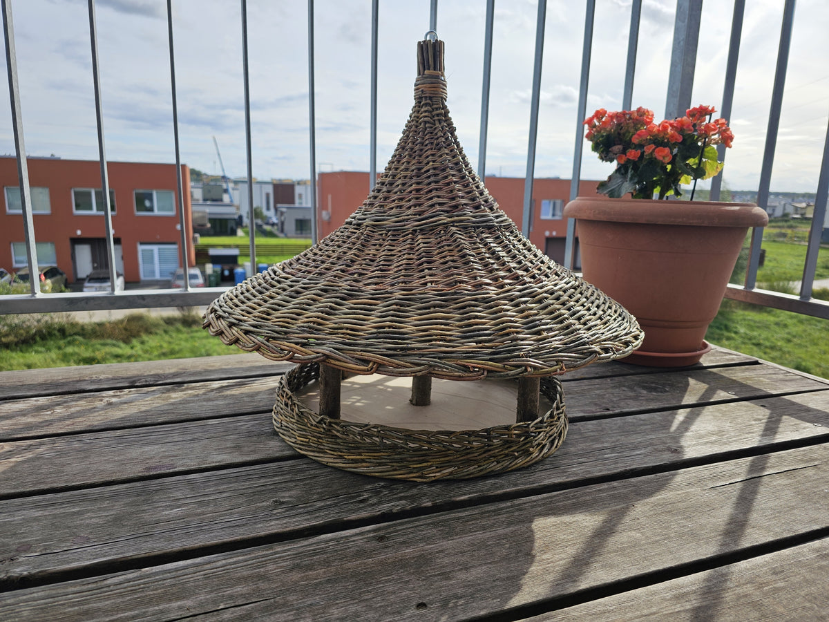Extra large wicker bird feeder standing on a wooden balcony table next to a flower pot, pointed roof and round woven base