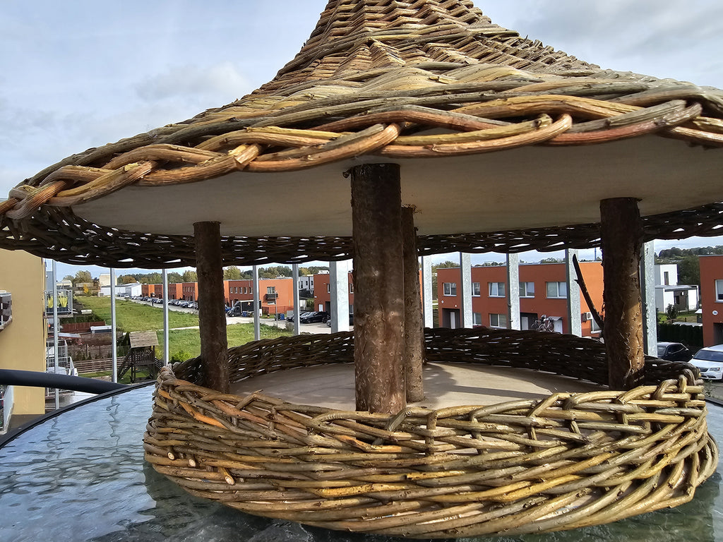 Low close-up of the bird feeder interior: branch posts holding the woven roof above the wooden platform and woven rim
