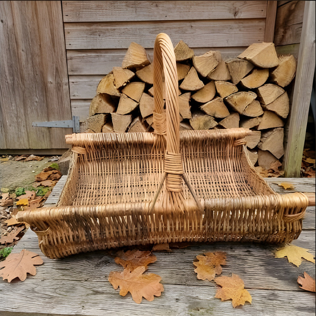 Large willow firewood basket with a tall handle on a wooden deck with autumn leaves, in front of a neatly stacked woodpile