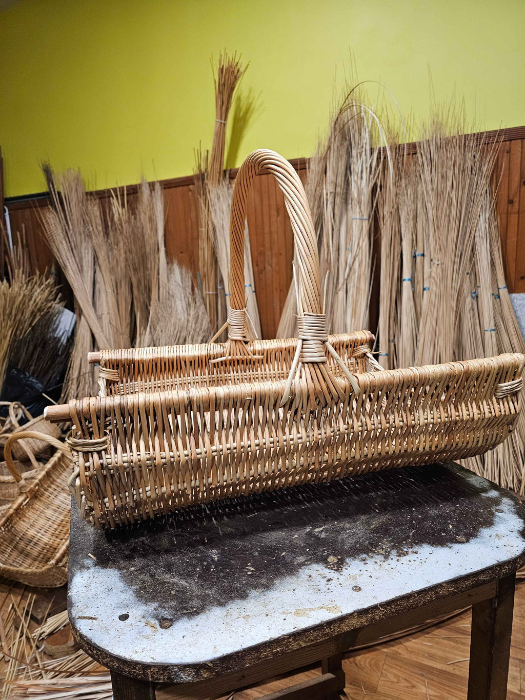Long handwoven willow log carrier with a tall twisted handle on a workbench in the weaving workshop, bundles of dried willow rods stacked against the wall.