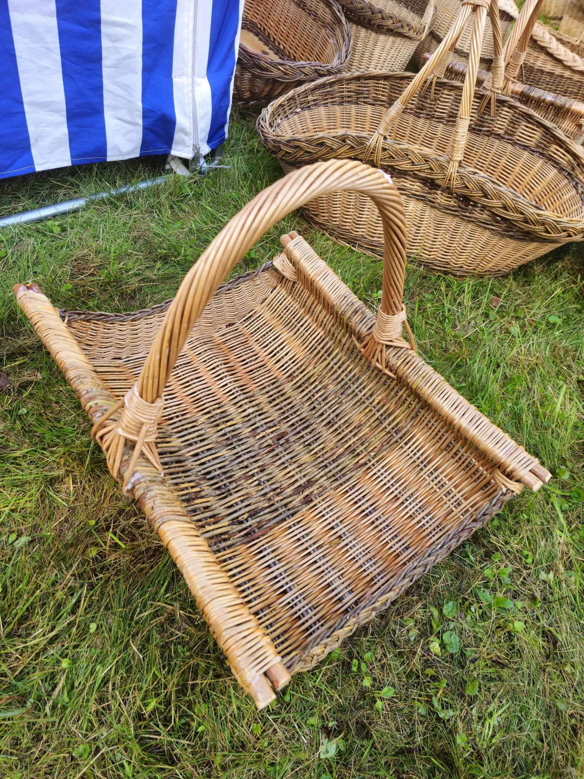 Group of handwoven willow baskets in different shapes on the grass, with a large rectangular log basket and its arched handle in the foreground.