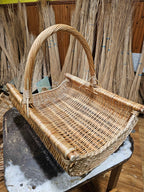 Rectangular handwoven willow log basket with a thick twisted handle standing on a workbench, surrounded by bundles of willow rods in the workshop.