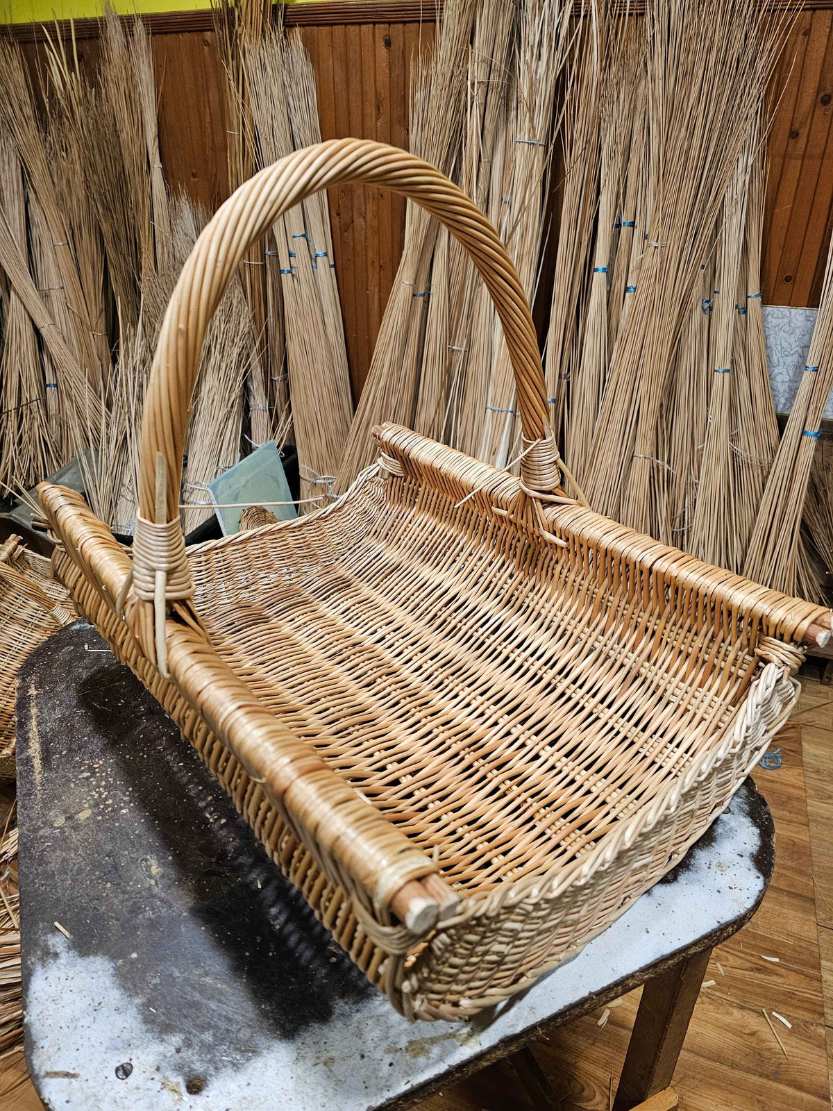 Rectangular handwoven willow log basket with a thick twisted handle standing on a workbench, surrounded by bundles of willow rods in the workshop.