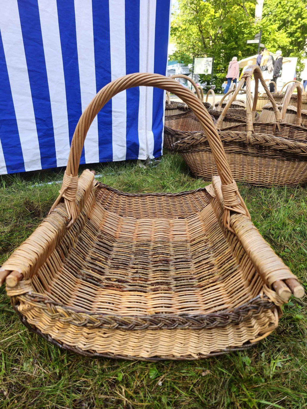 Large handwoven wicker firewood trug with curved sides, thick side rods and a tightly wound arched handle on grass at a craft fair