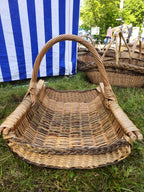 Large handwoven wicker firewood trug with curved sides, thick side rods and a tightly wound arched handle on grass at a craft fair