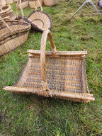Rectangular handwoven willow firewood basket with a curved handle and thick side rods on the grass, with other wicker baskets around it