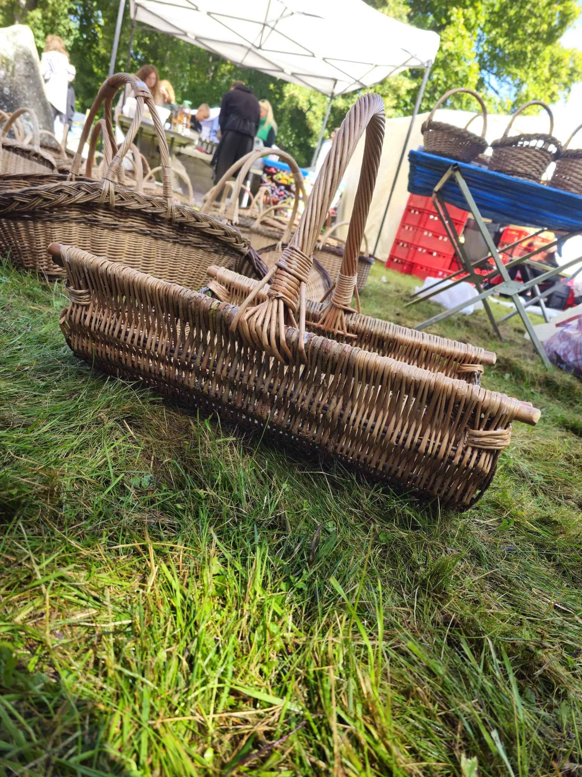 Willow log carrier with a tall handle standing on the grass at an outdoor craft market, with more handwoven baskets and stalls behind