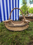 Large handwoven willow log basket with a curved handle, thick side rods and a woven border, set on the grass in front of a striped tent