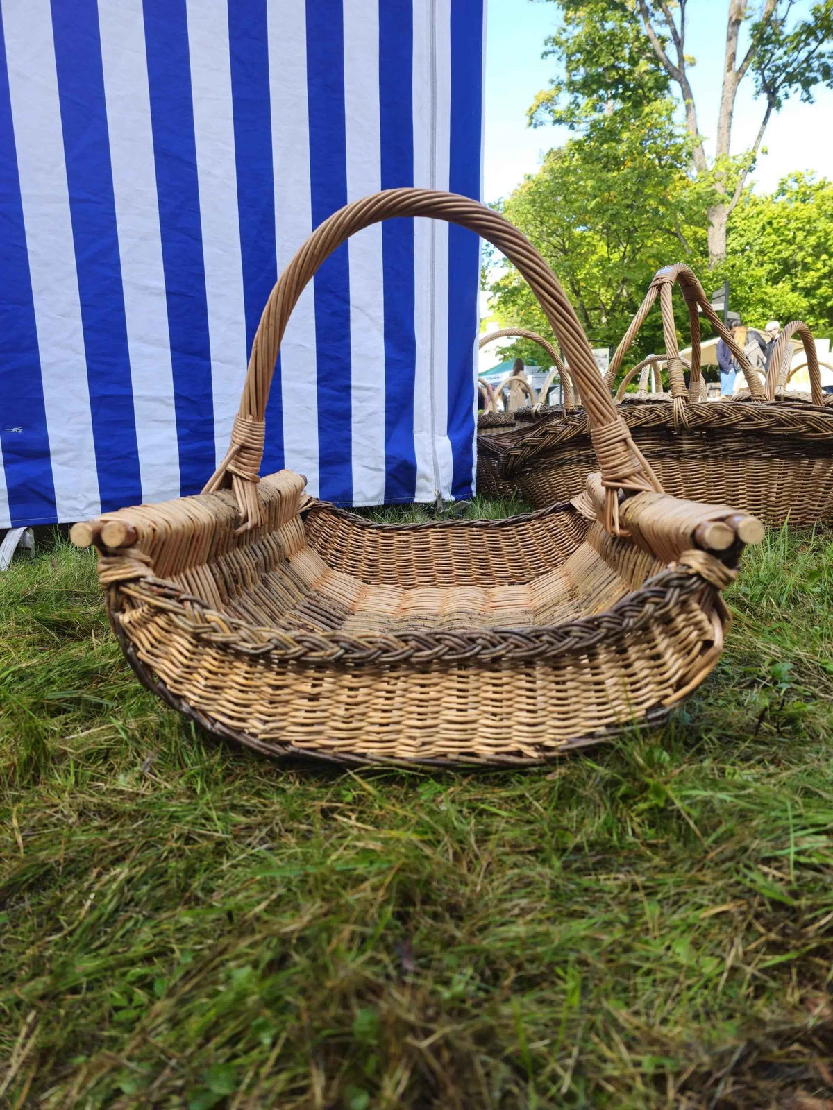 Large handwoven willow log basket with a curved handle, thick side rods and a woven border, set on the grass in front of a striped tent