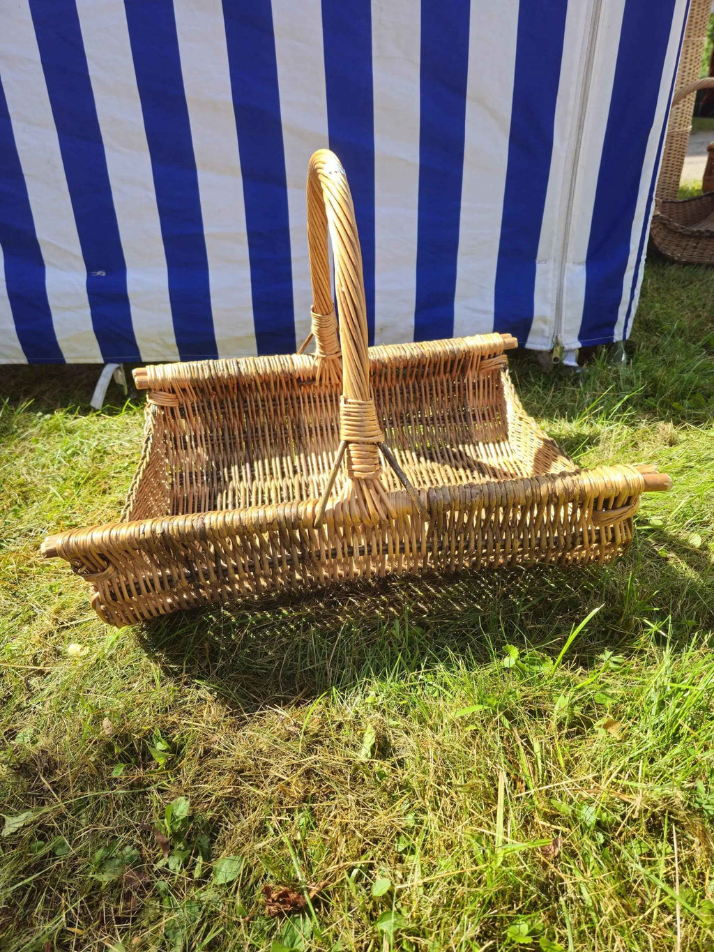 Rectangular handwoven willow firewood basket with a tall curved handle and low sides, standing on green grass in the sunshine