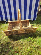 Rectangular handwoven willow firewood basket with a tall curved handle and low sides, standing on green grass in the sunshine