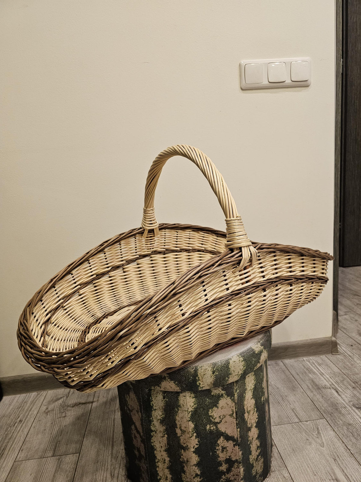 Elongated handwoven willow garden trug with a high arched handle displayed on a round stool indoors.