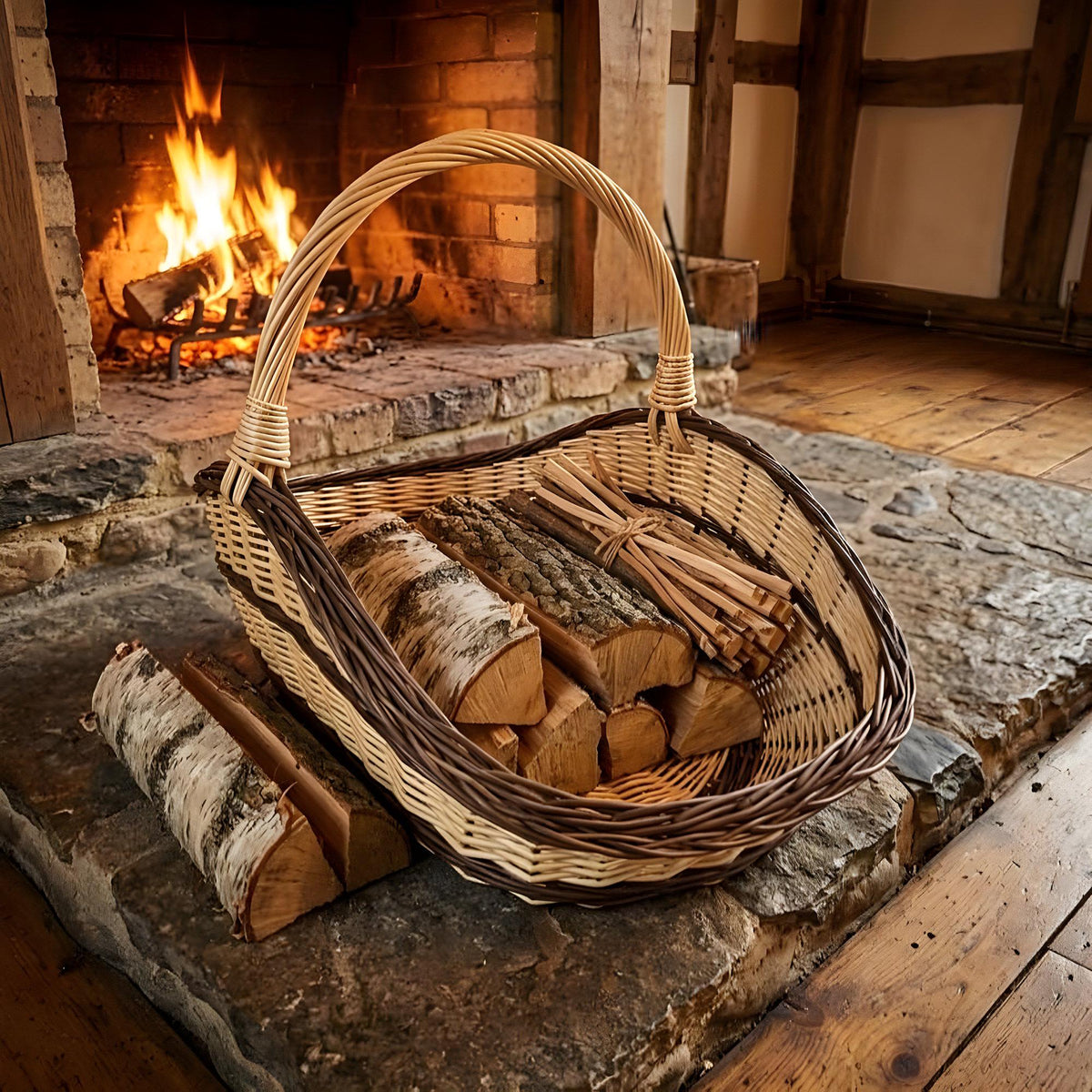 Large willow trug basket filled with birch logs and kindling on the hearth in front of a burning fireplace.