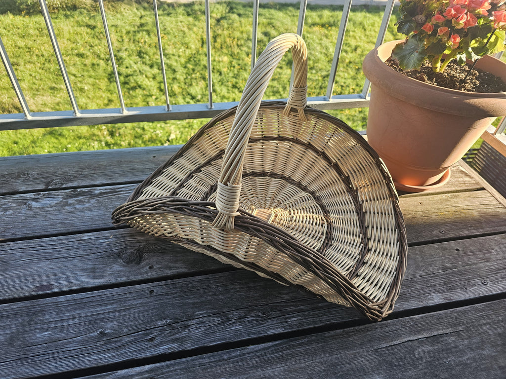 Large willow flower gathering trug with tall handle on a wooden balcony table beside a pot of flowers