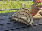 Large willow flower gathering trug with tall handle on a wooden balcony table beside a pot of flowers