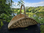 Large boat-shaped wicker basket with an arched handle standing on dark ground above a river valley, its open curved body facing the view.