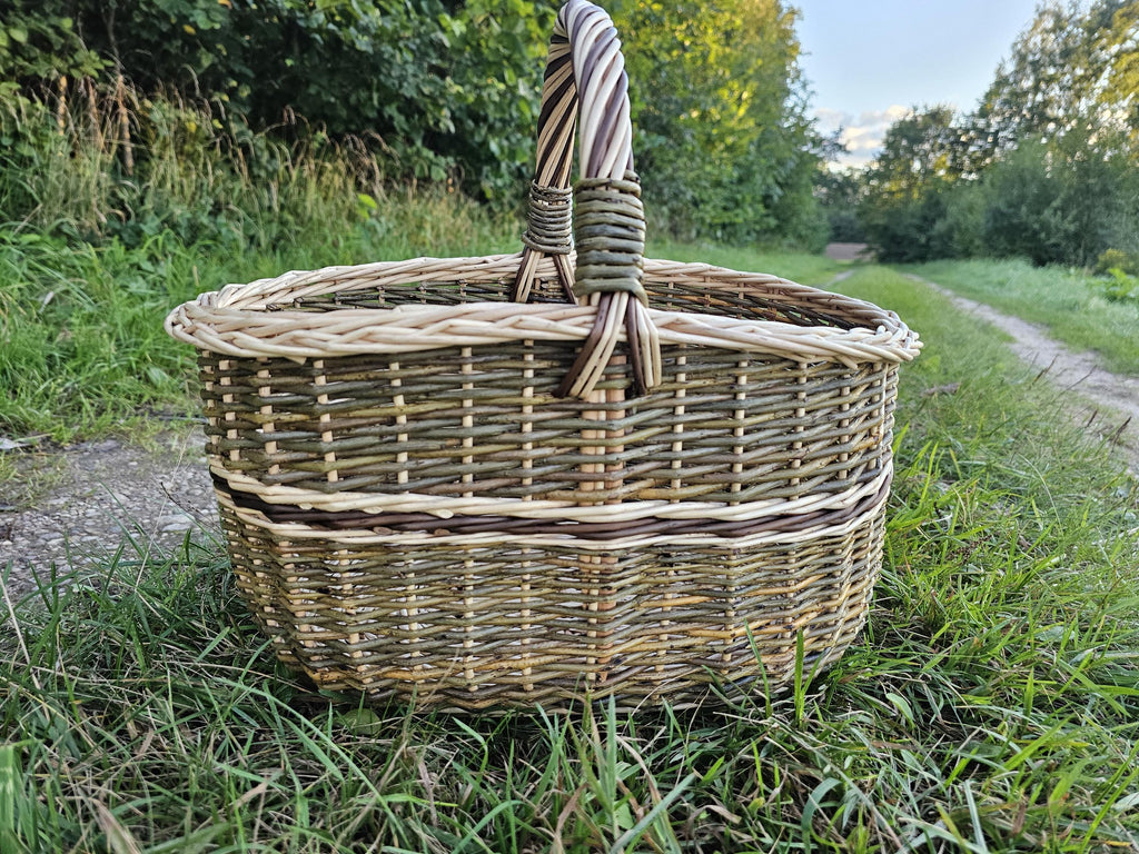 Side view of the oval wicker market basket with its twisted handle, standing in tall grass beside a country path