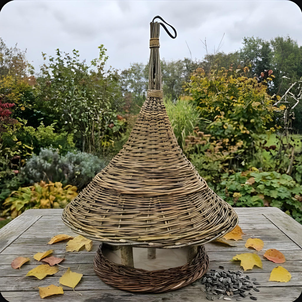 Handwoven willow bird feeder with a tall conical roof and hanging loop on a garden table with autumn leaves and sunflower seeds