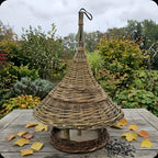 Handwoven willow bird feeder with a tall conical roof and hanging loop on a garden table with autumn leaves and sunflower seeds