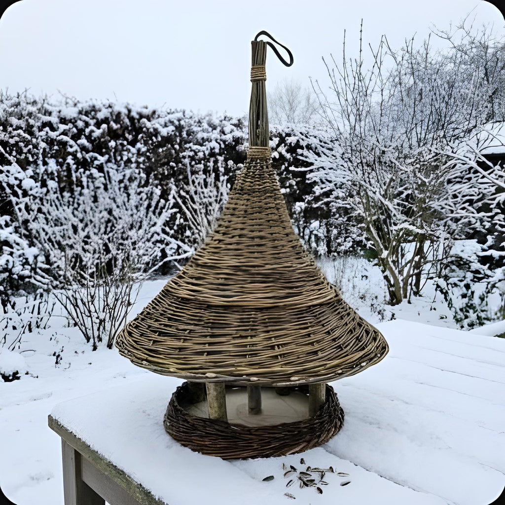 Hanging willow bird feeder with a conical roof on a snow-covered garden table in winter, with sunflower seeds scattered in the snow