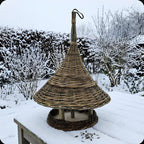 Hanging willow bird feeder with a conical roof on a snow-covered garden table in winter, with sunflower seeds scattered in the snow